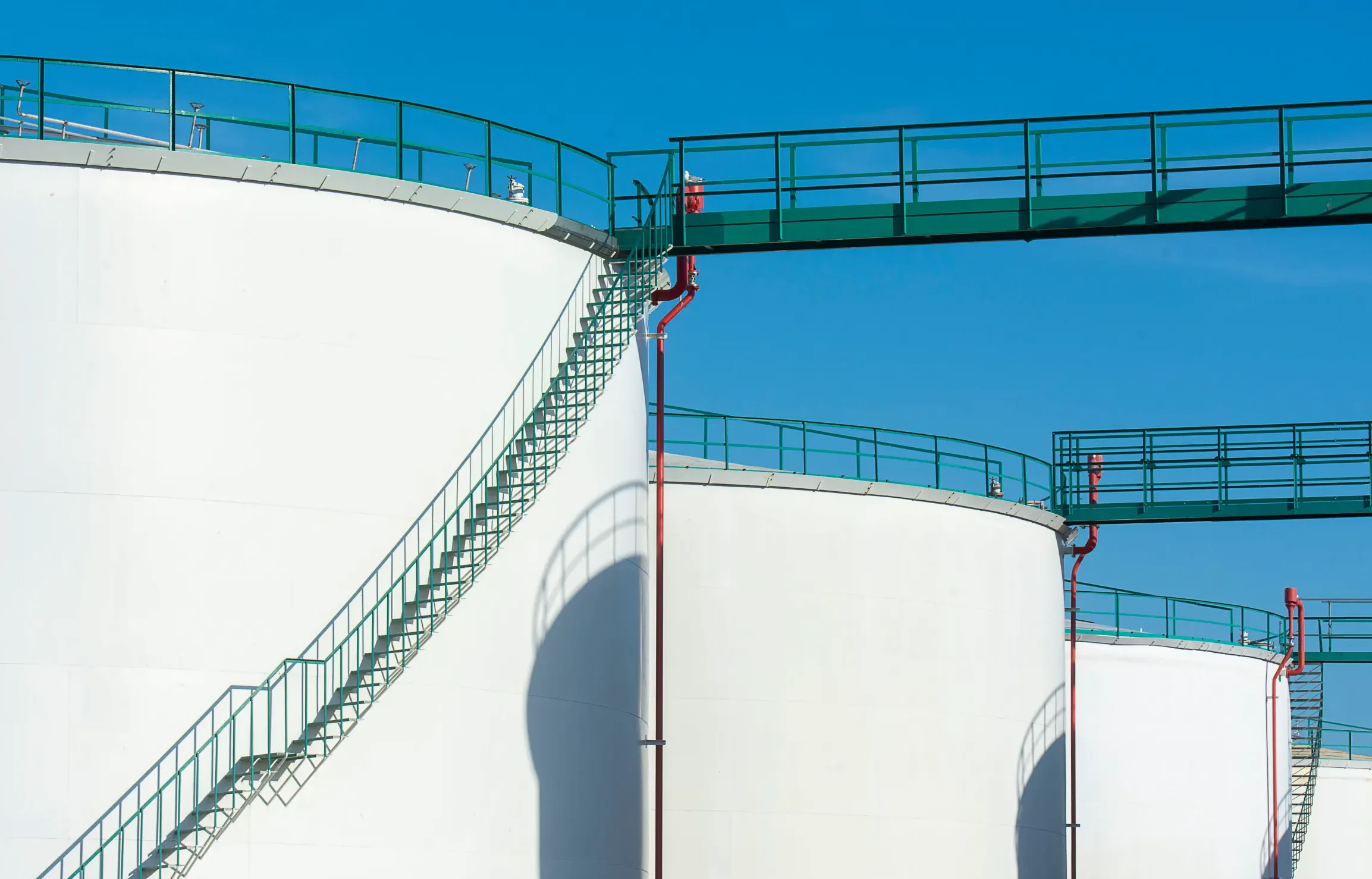 White storage tanks joined by green gantries and stair rails, against a clear blue sky.